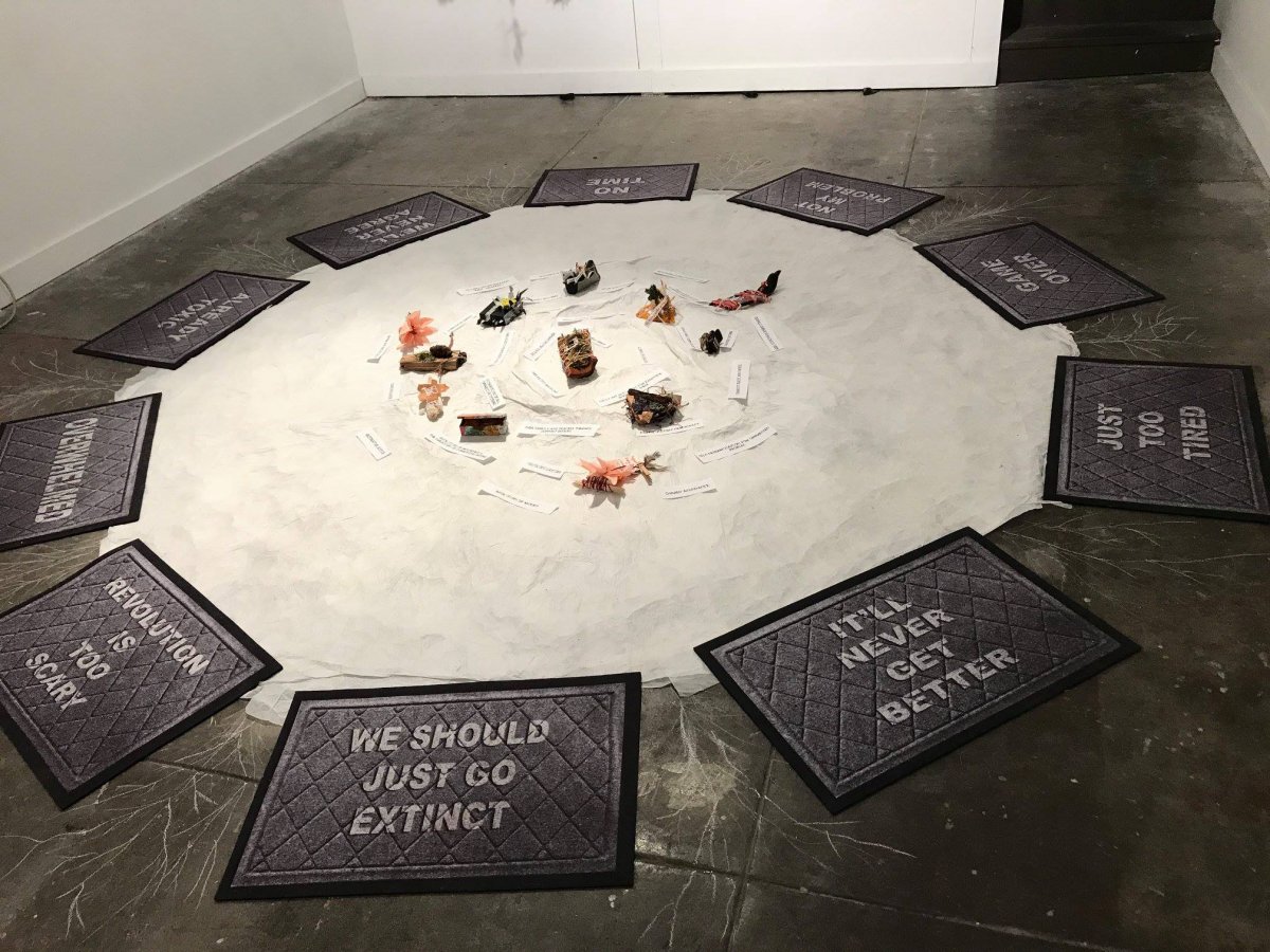 Beverly Naidus: We Almost Didn't Make It A white circle of material on the floor is surrounded by mats with messages printed on such as 'Revolution is too scary', ' We should just go extinct', etc. At the centre are some small objects which appear to be made of shells and other natural materials