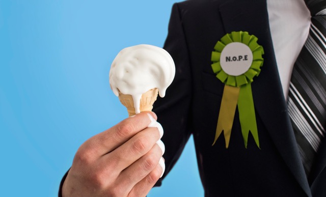 photo by Jamie Rivers An ice cream is held in the hand of a person wearing a suit jacket, on their jacket is a rosette with the letters N.O.P.E.