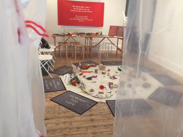 A gallery space contains a white floor mat covered in objects and texts, surrounded by door mats with texts including 'Already Too Late' and 'How Can I Give Hope to a Child?'