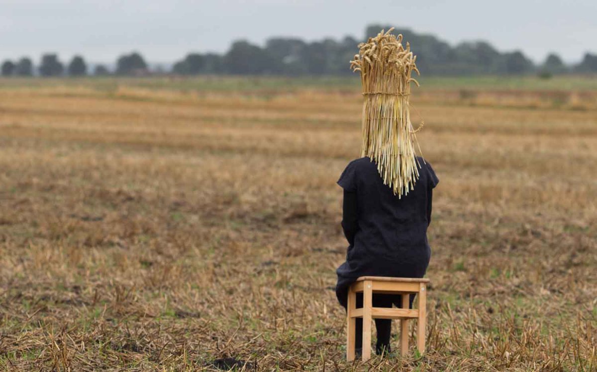 Image: Anne-Marie Culhane In a harvested field a figure sites on a wooden stool with their head covered by a sheath of corn.