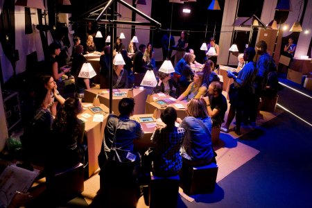 Audience members/participants in a performance gather around cardboard tables, with discussion points written on paper lamps above them.